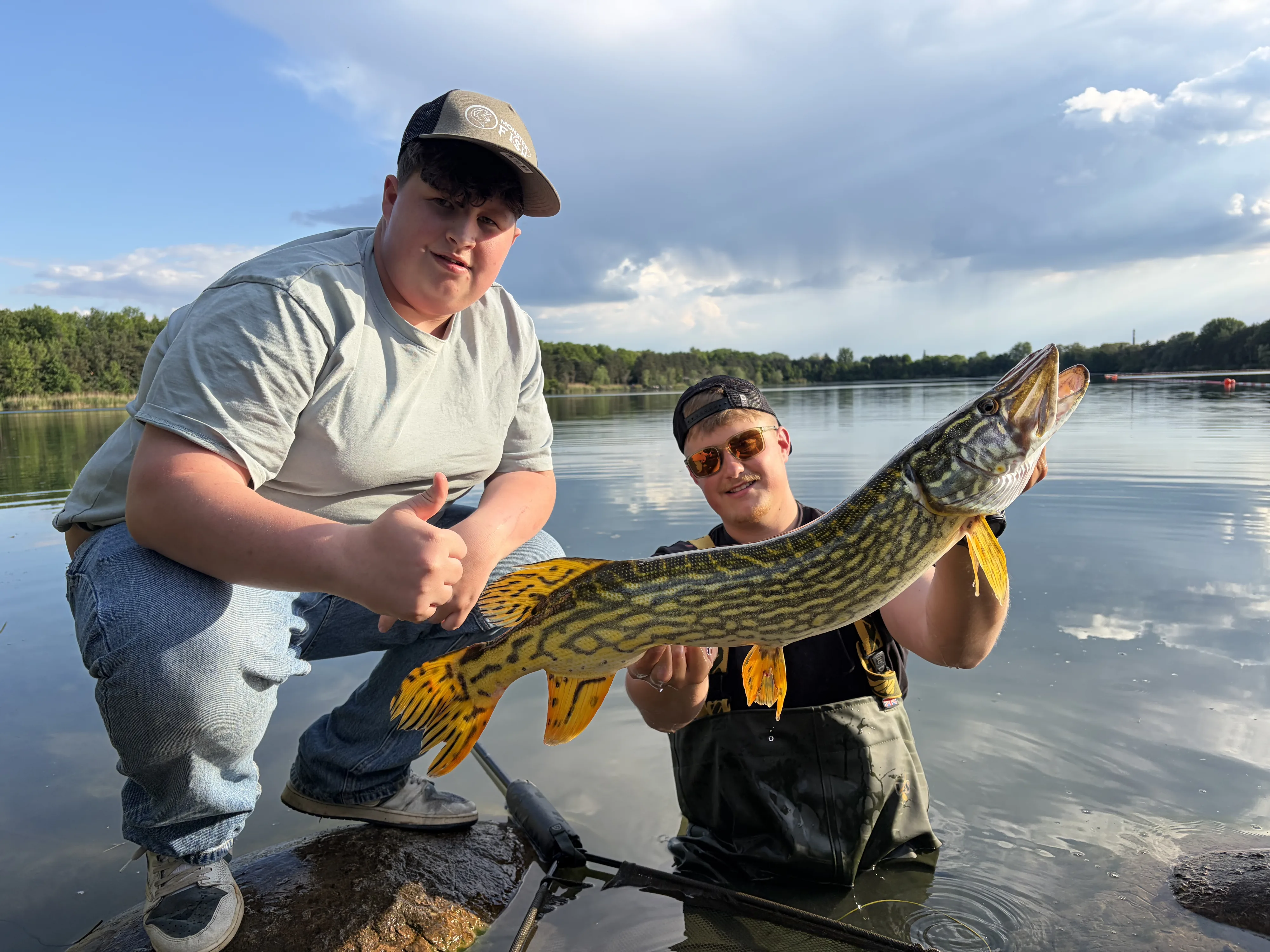 Zwei Angler am Ufer – einer steht im flachen Wasser und hält einen kräftig gefärbten Hecht