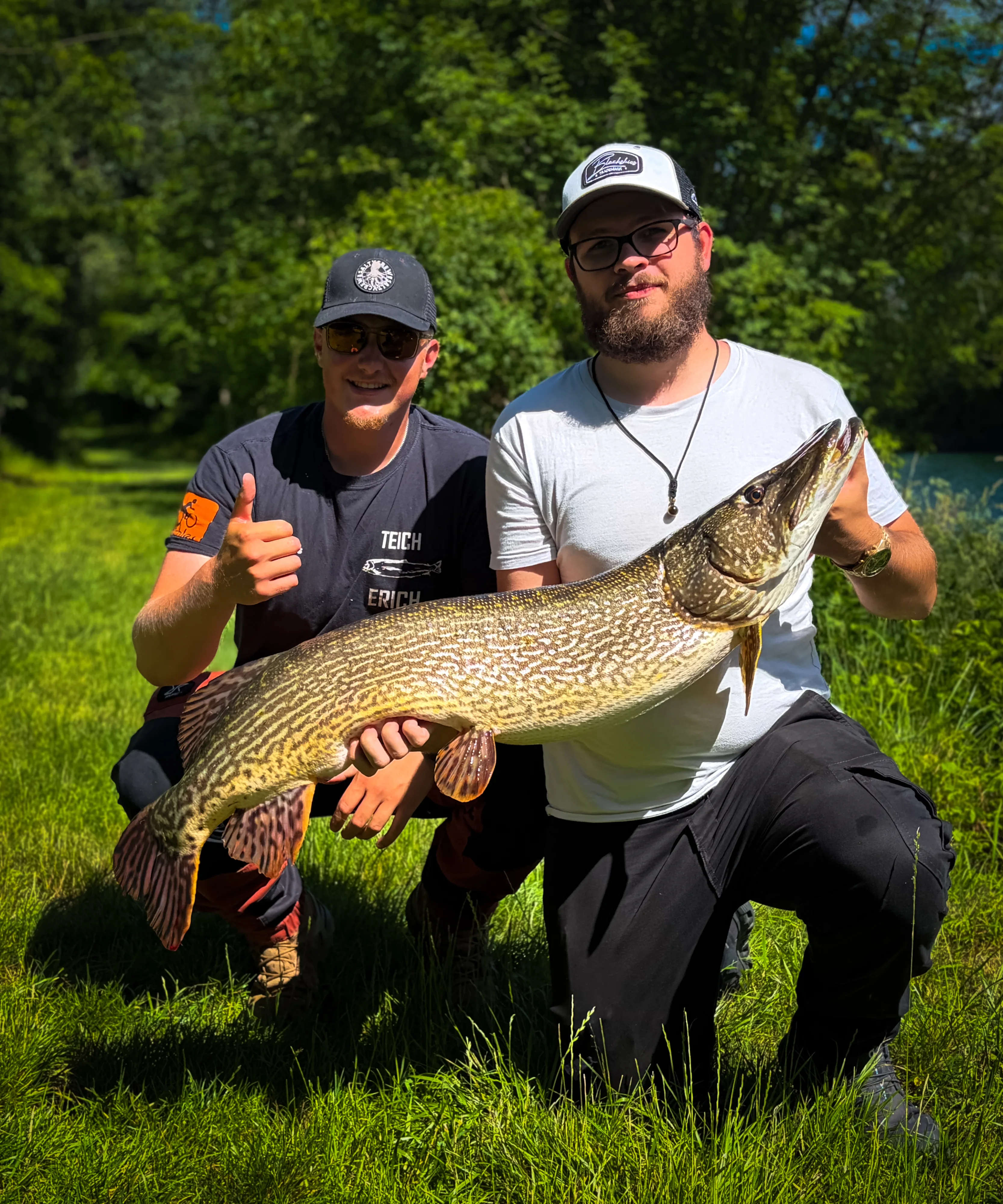 Zwei Angler knien auf der Wiese mit einem sehr großen Hecht
